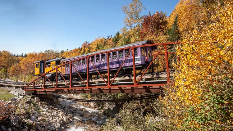 An orange and purple train crosses over a small but rapid river, against a backdrop of vivid foliage.